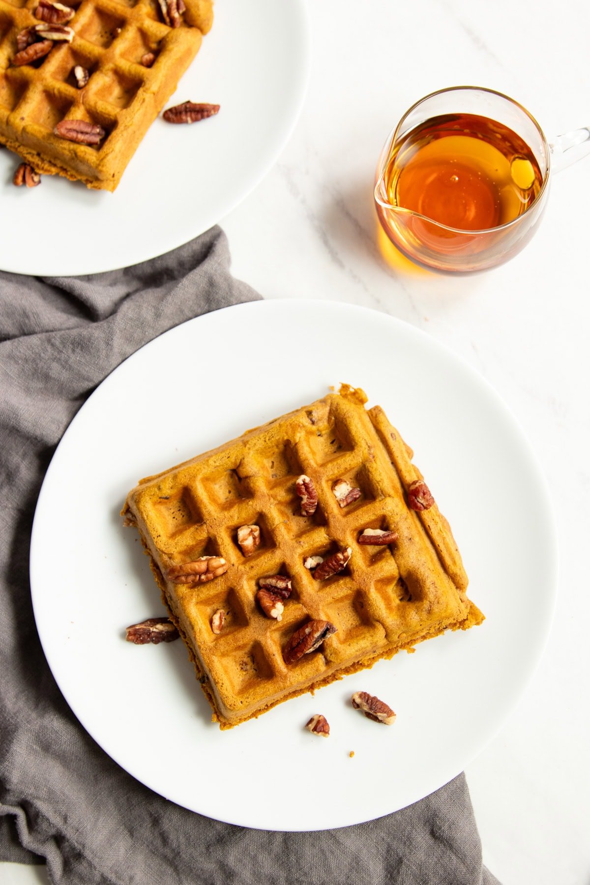 Looking down at two plates each containing one pumpkin waffle topped with chopped pecans. A small glass jar of maple syrup sits off to the side. 