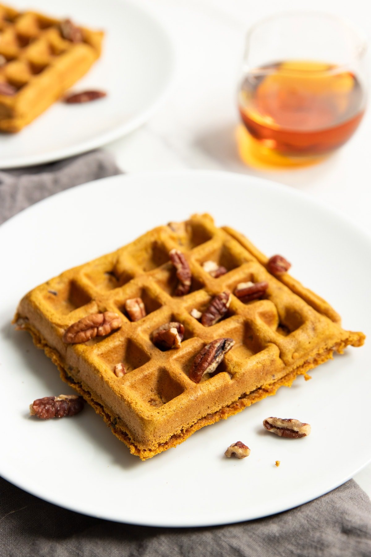 Two plates each containing a square pumpkin spice waffle are staggered on counter. There's a small glass pitcher of maple syrup in. between. 
