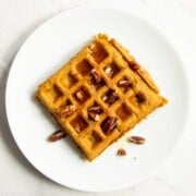 Looking down at a smooth white plate holding a naturally orange-colored, square waffle topped with chopped pecans and maple syrup.