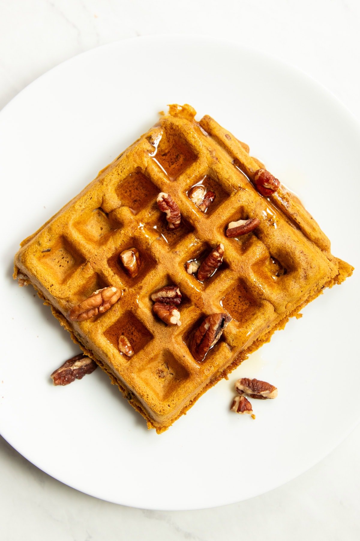 A low FODMAP pumpkin spice waffle topped with chopped pecans and maple syrup on a white plate.