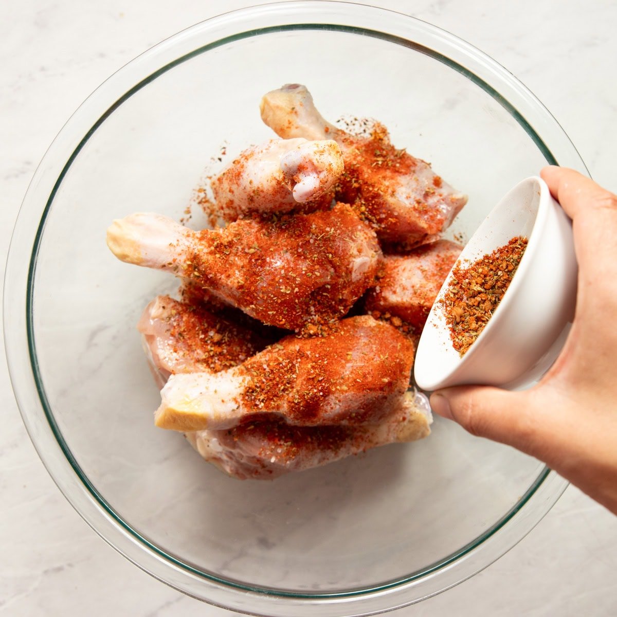 Sprinkling a homemade seasoning on top of oil coated chicken drumsticks in a large mixing bowl.