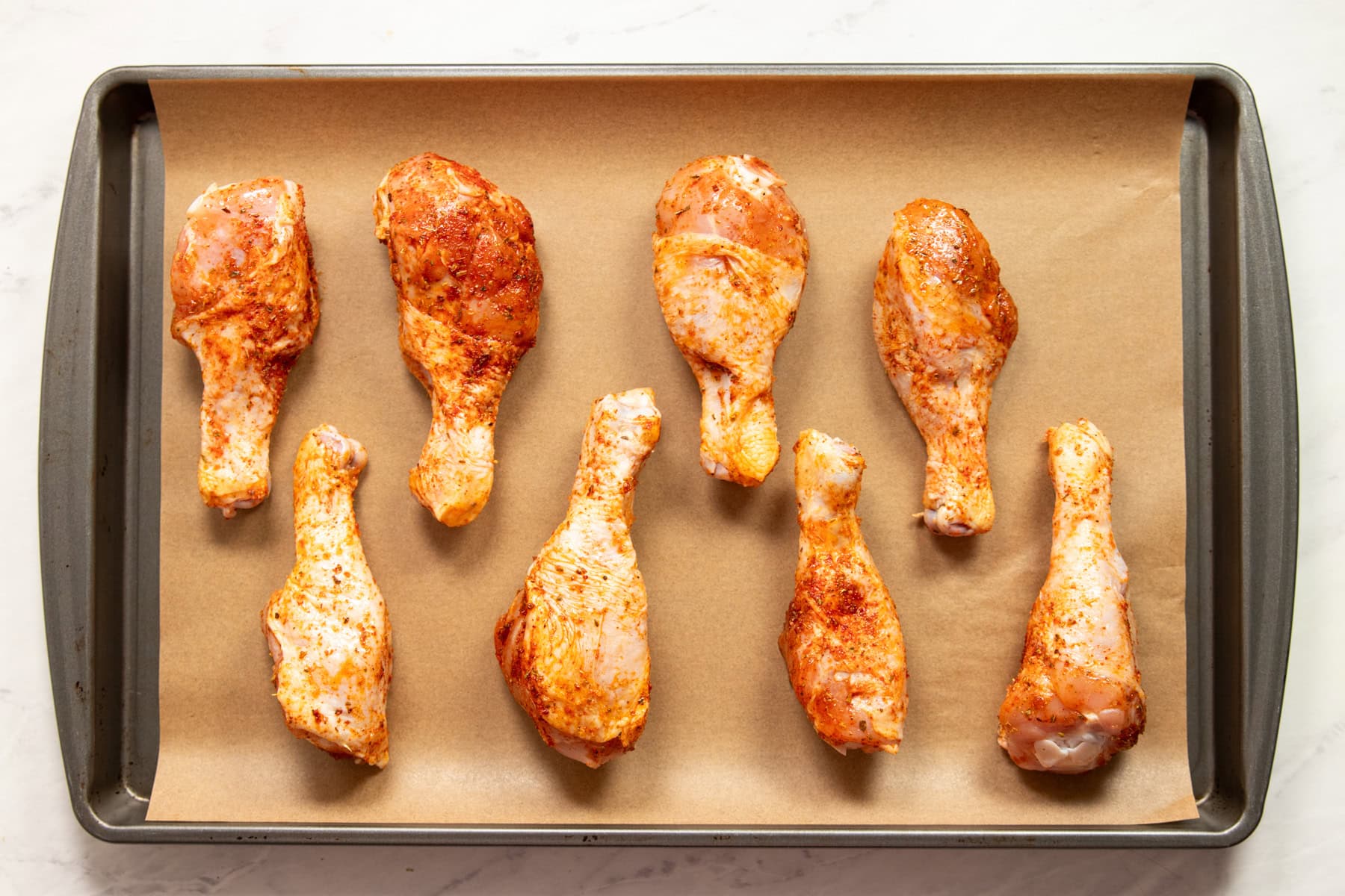 Oiled and seasoned chicken drumsticks are spread out onto a large baking sheet lined with parchment paper ready for baking.