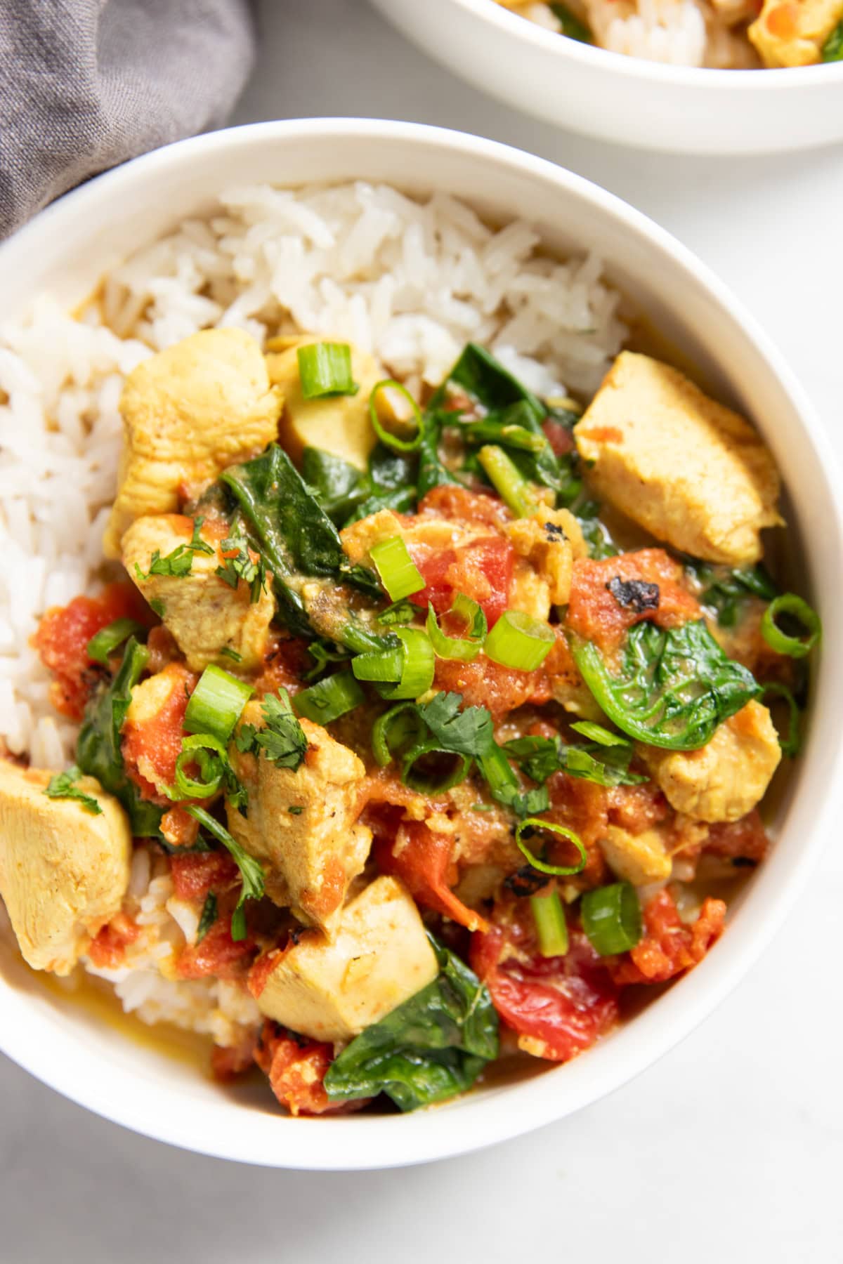 A close up of chicken curry over white rice. There are chunks of tender cooked chicken with diced tomatoes, wilted spinach, fresh herbs and yellow curry sauce.