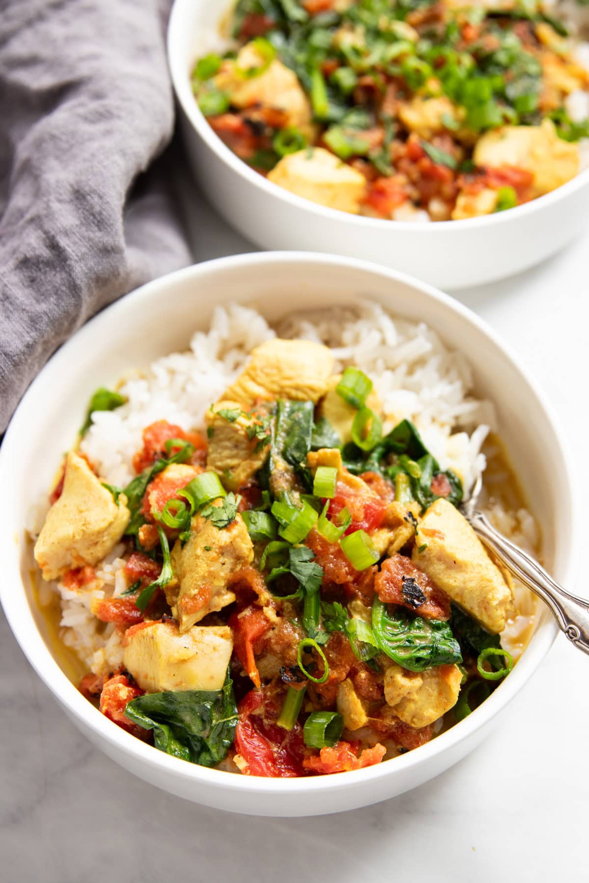 Two bowls are staggered on a white counter with a gray linen napkin in between. Each bowl contains chicken curry with tomatoes and spinach over white rice. A spoon rests in one of the bowls ready to eat.