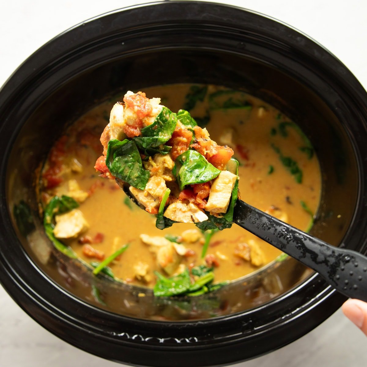 A serving spoon holds a scoop of chicken curry over the slow cooker.