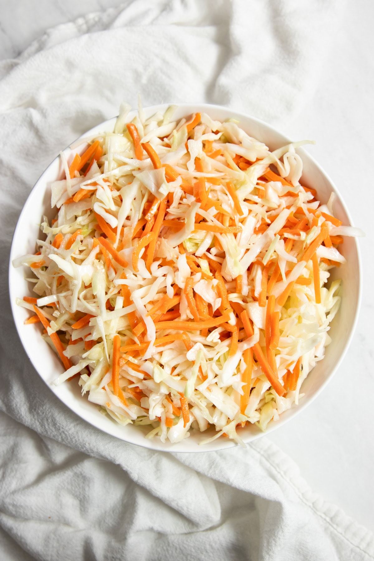 A bowl of low FODMAP vinegar coleslaw sits on a white counter with a white cotton napkin artistically surrounding the bowl. 