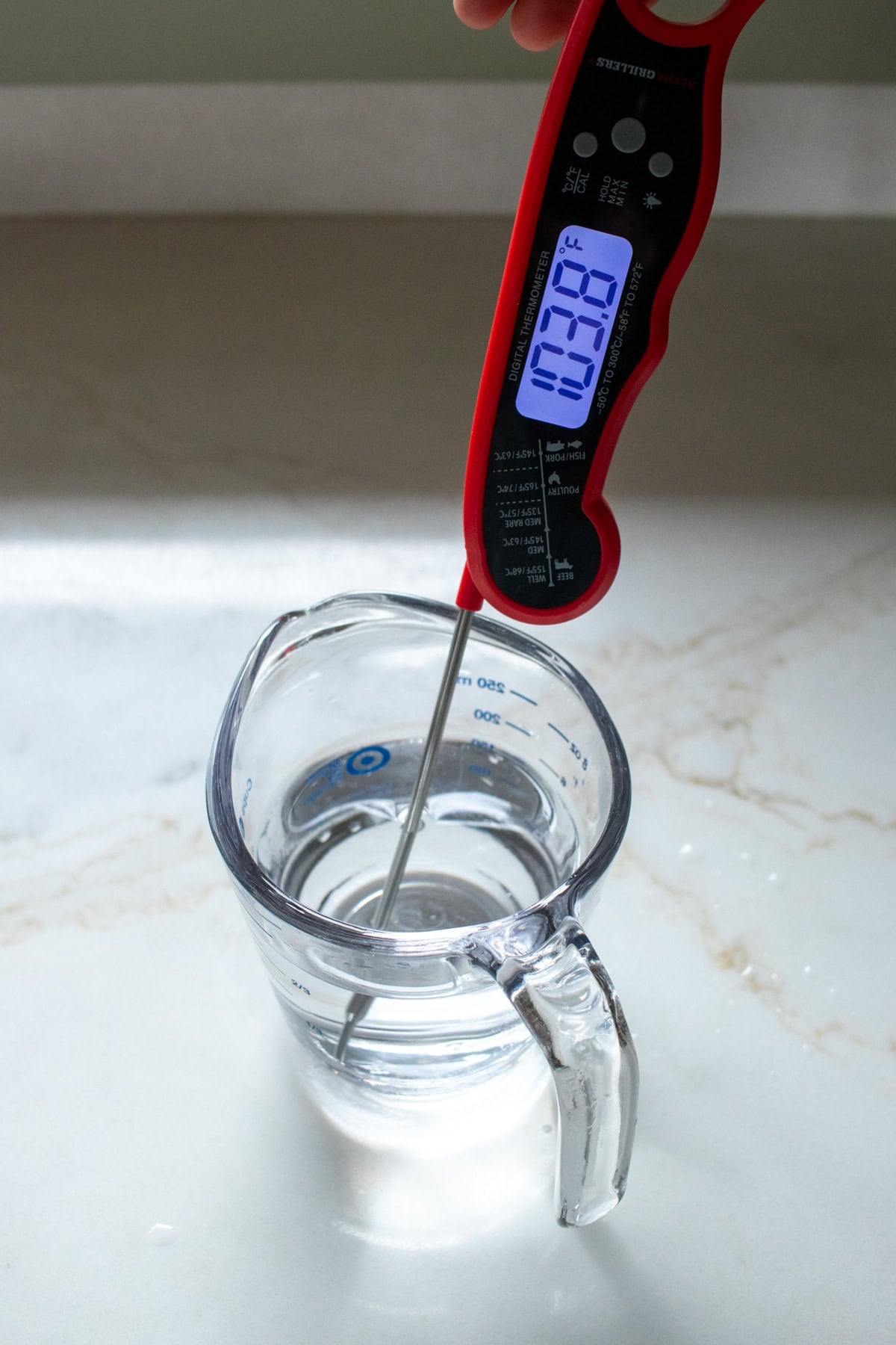 A food thermometer measuring the temperature of a glass measuring cup filled with water. The thermometer reads 103.8F.
