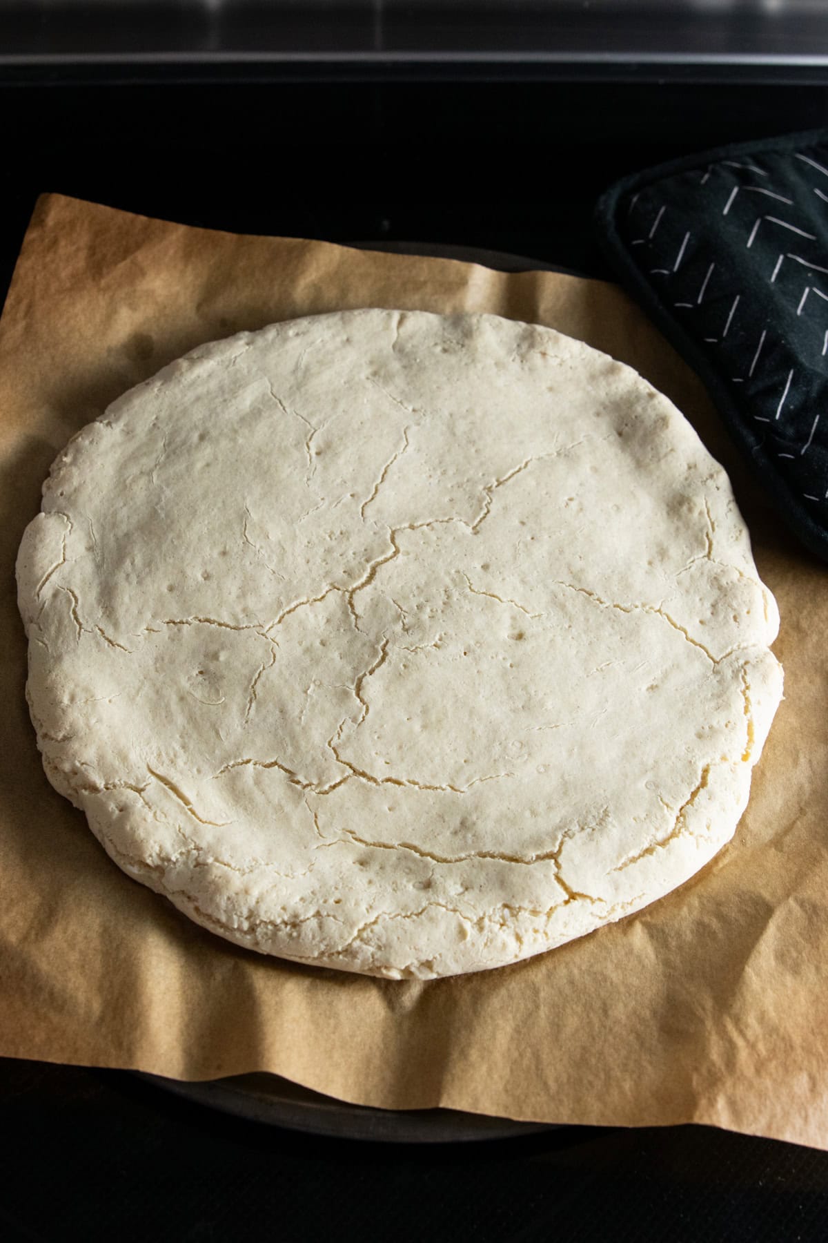 A par-baked low FODMAP pizza crust sitting on a parchment-lined pizza pan with a pot holder resting off to the side.