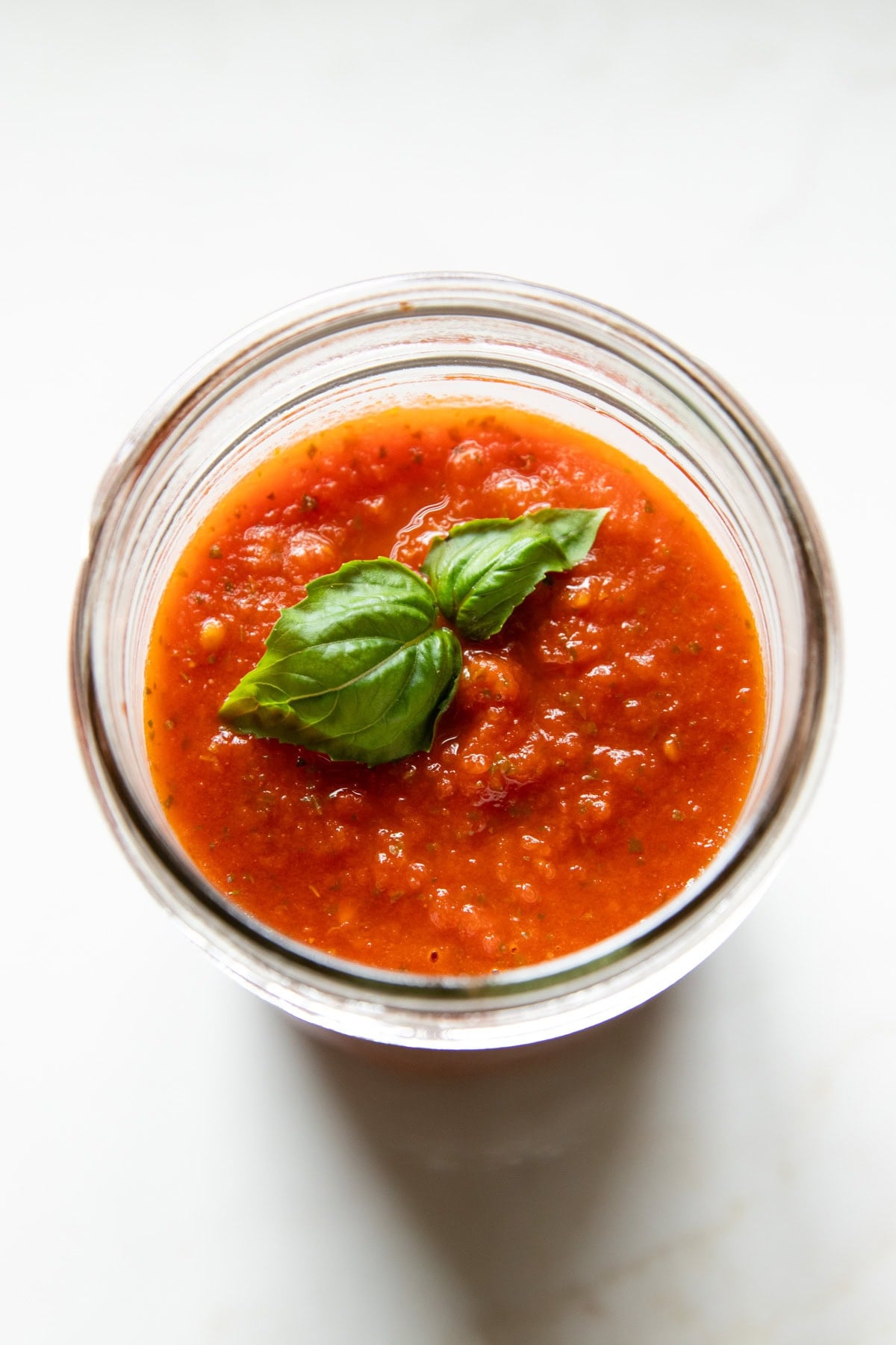 A glass jar of pizza sauce sits on white countertop. There are basil leaves sitting on top of the sauce.