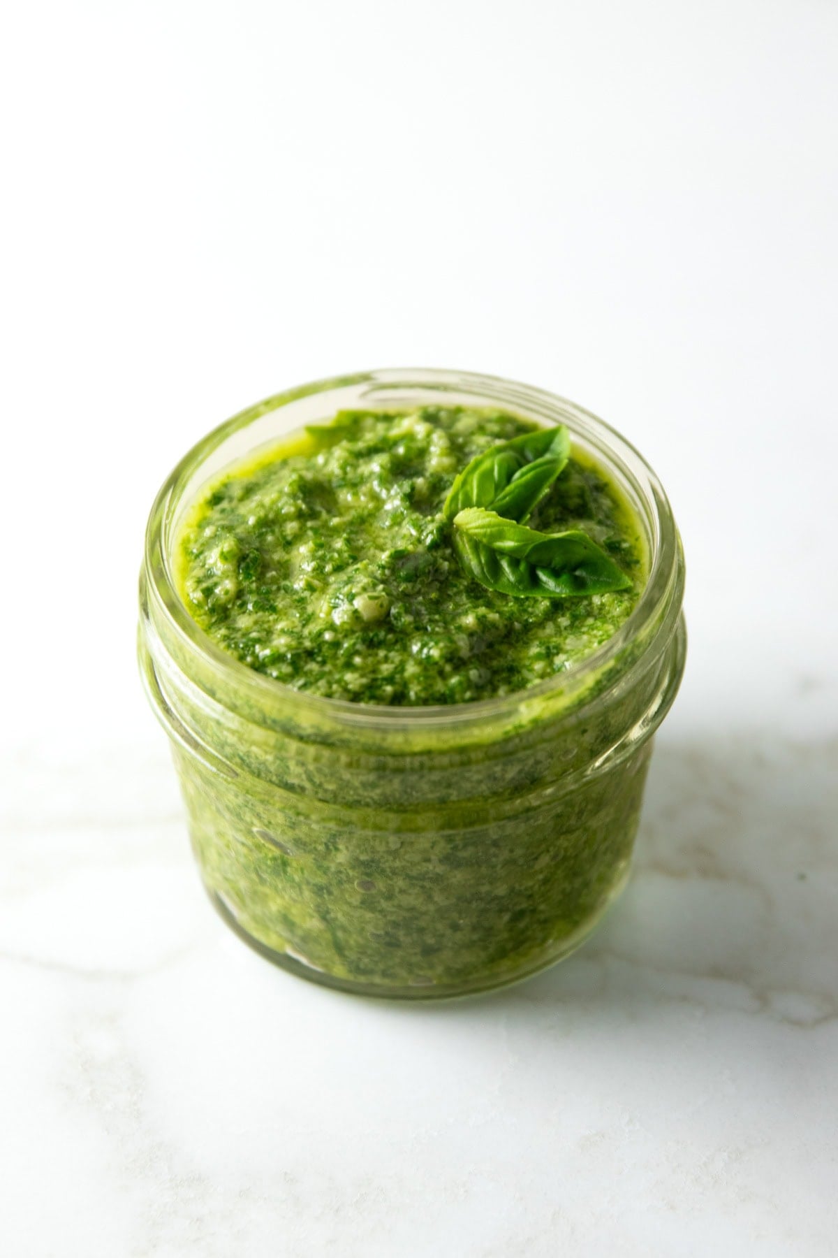 Small glass jar filled with vibrant green low FODMAP pesto, topped with a fresh basil sprig, set on a white counter.