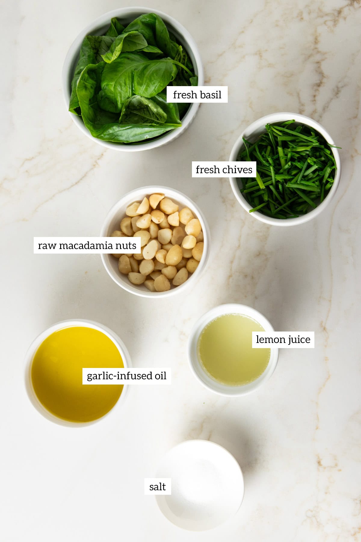 Ingredients for low FODMAP pesto in individual white dishes: fresh basil, chives, macadamia nuts, garlic-infused oil, lemon juice, and salt.