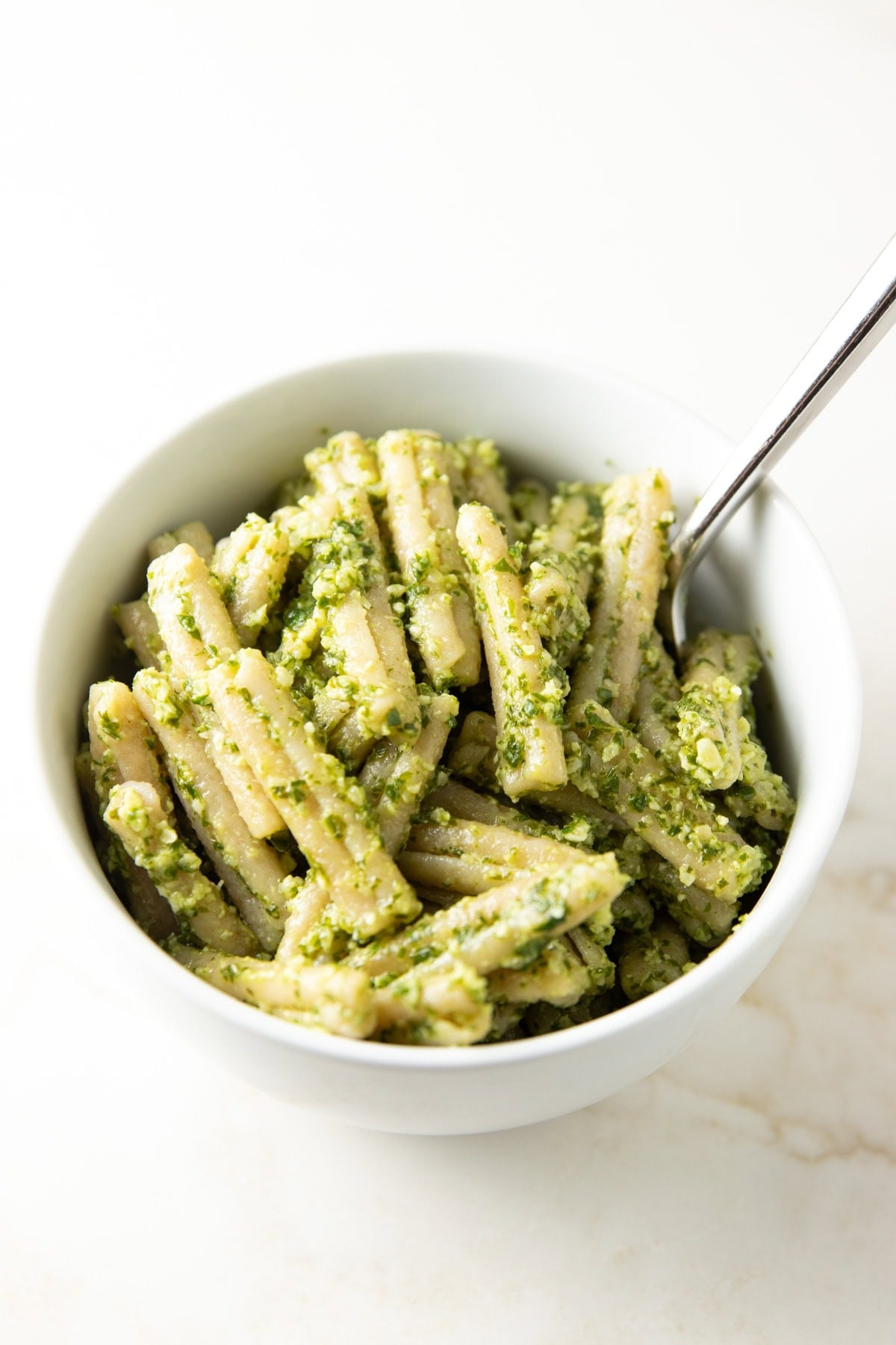 Bowl of brown rice pasta tossed in a chunky green pesto, coated evenly with visible bits of herbs and nuts, with a spoon resting on the side.