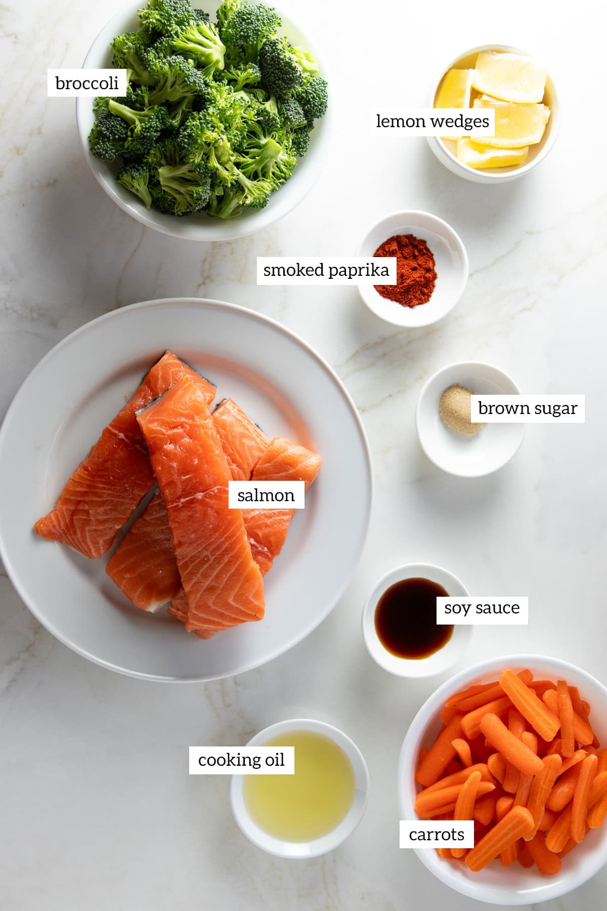 Ingredients for sheet pan salmon including raw salmon fillets, broccoli florets, baby carrots, lemon wedges, smoked paprika, brown sugar, soy sauce, and cooking oil arranged in bowls on a white counter.