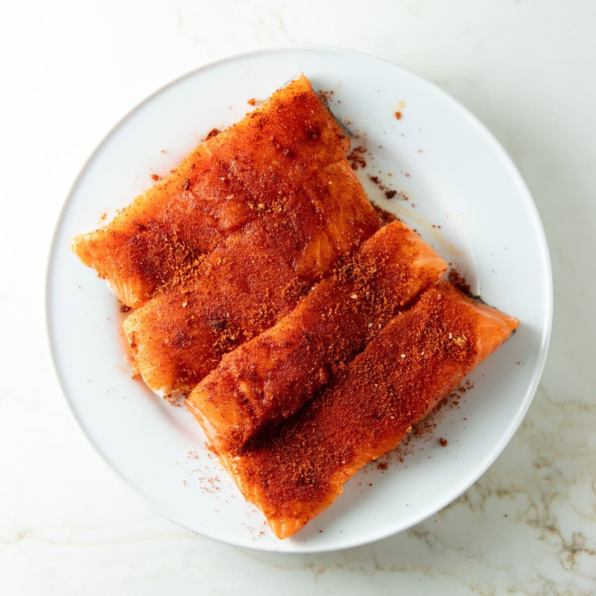 Four salmon fillets coated with smoked paprika, brown sugar, and salt arranged on a white plate