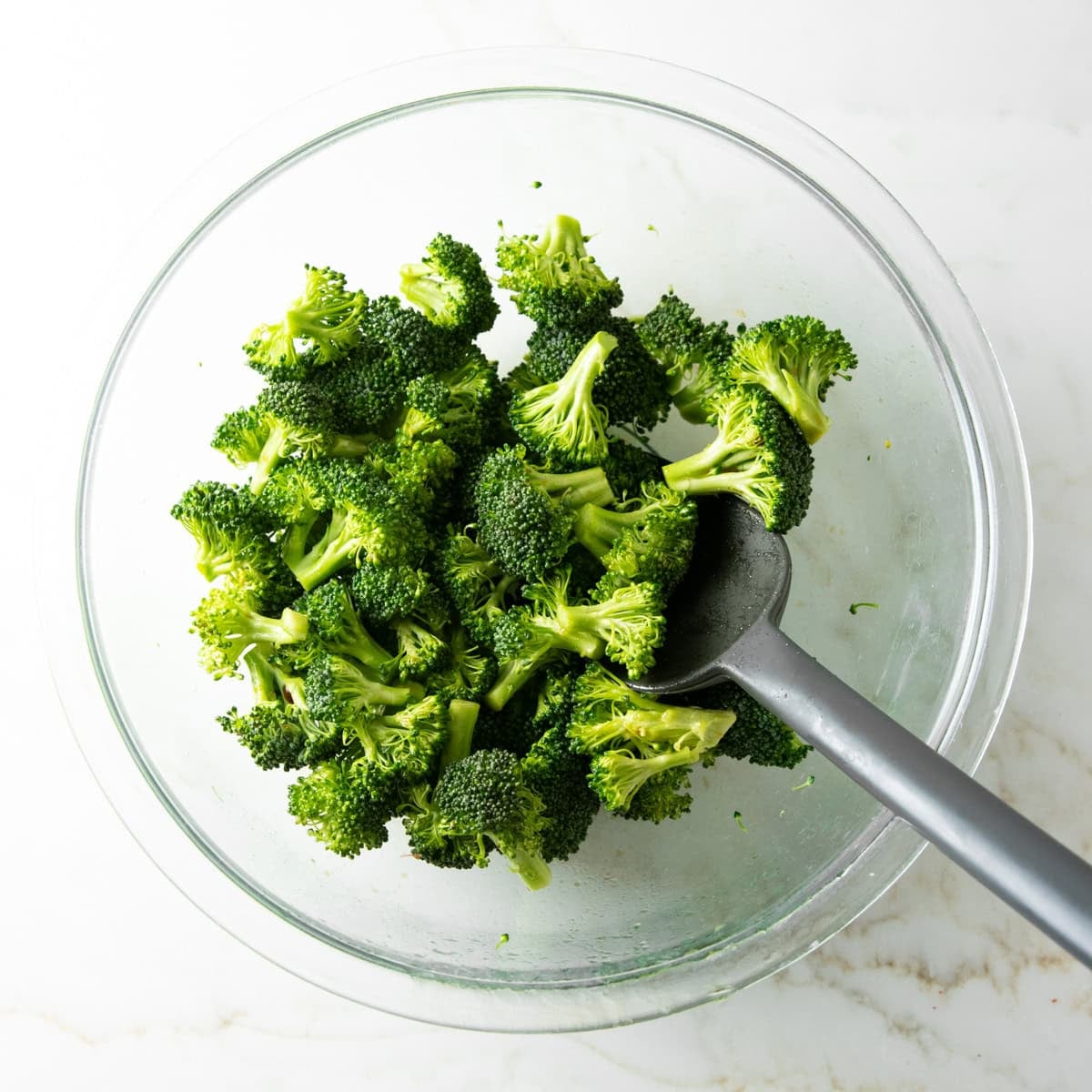 Broccoli florets tossed with oil and soy sauce in a bowl using a silicone spatula