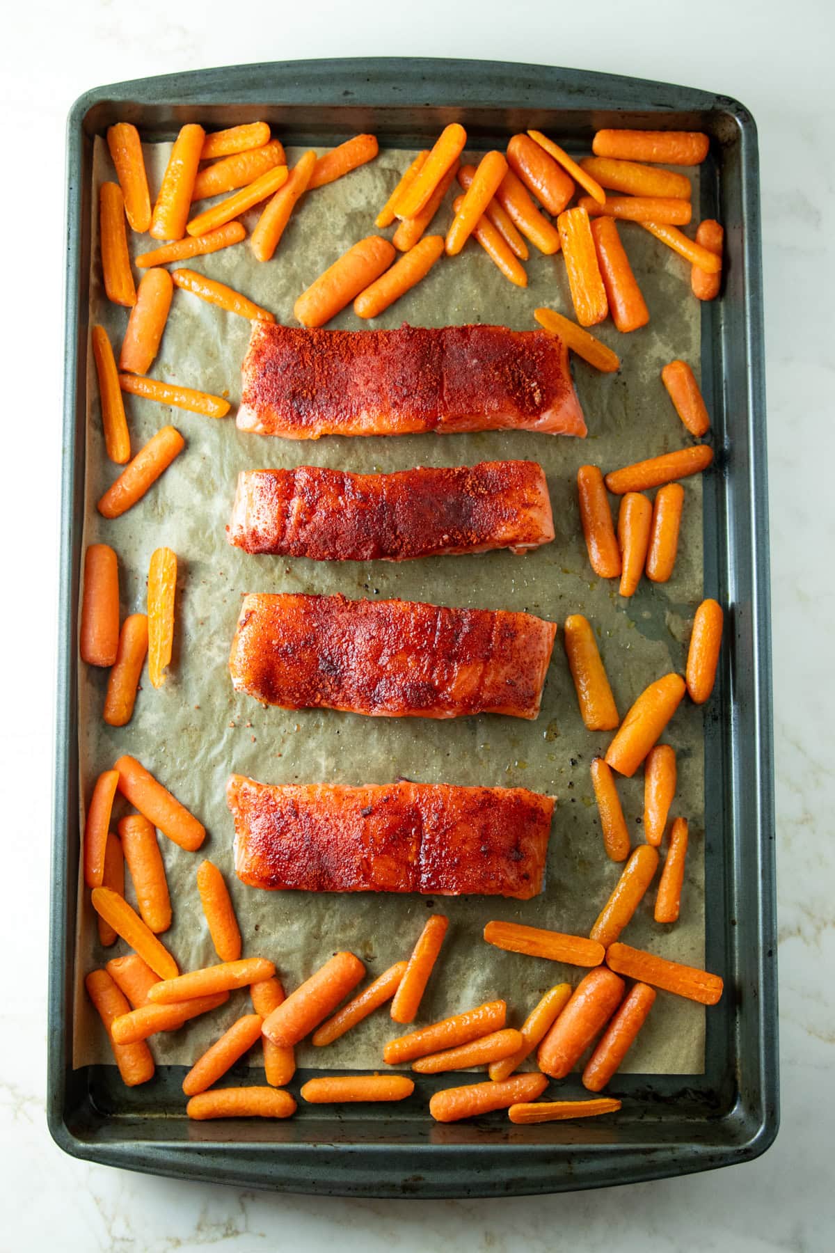 Salmon fillets with a smoked paprika-brown sugar rub arranged in the center of a parchment-lined sheet pan, surrounded by partially roasted baby carrots.