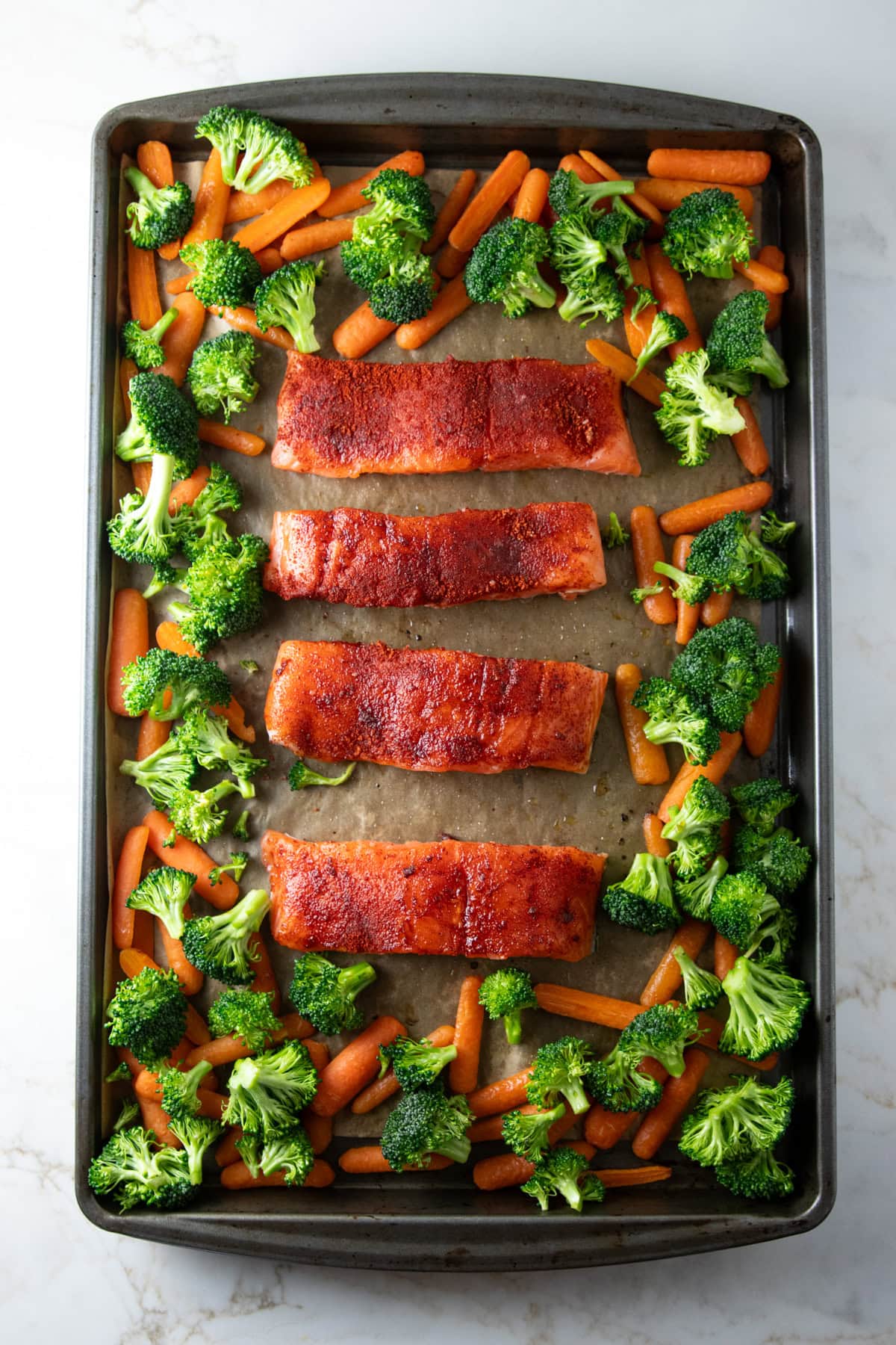 Salmon fillets with a smoked paprika rub arranged in the center of a parchment-lined sheet pan, surrounded by partially roasted baby carrots. Broccoli florets are sprinkled on top of the carrots ready for roasting.