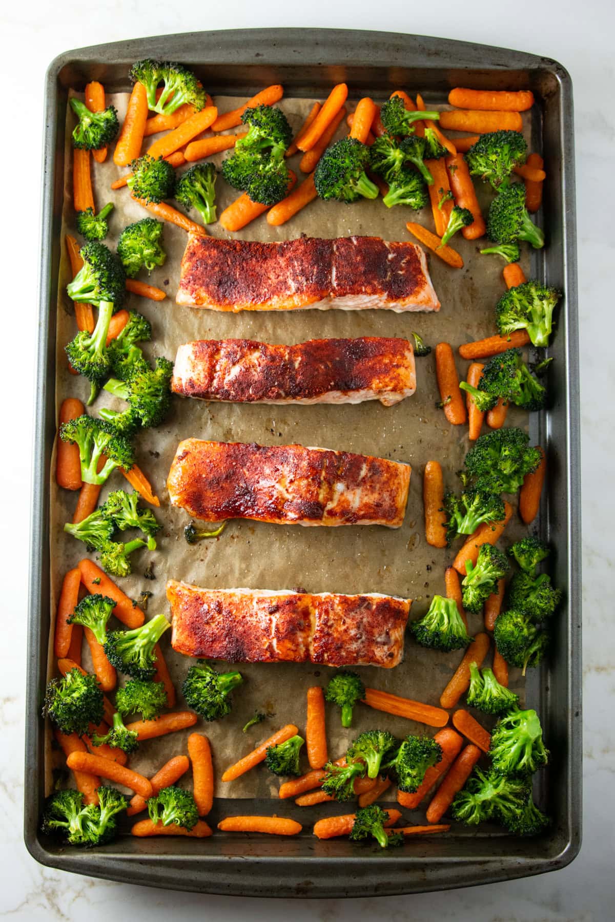 Sheet pan baked salmon fillets with roasted broccoli florets and baby carrots arranged around the edges on a parchment-lined baking sheet
