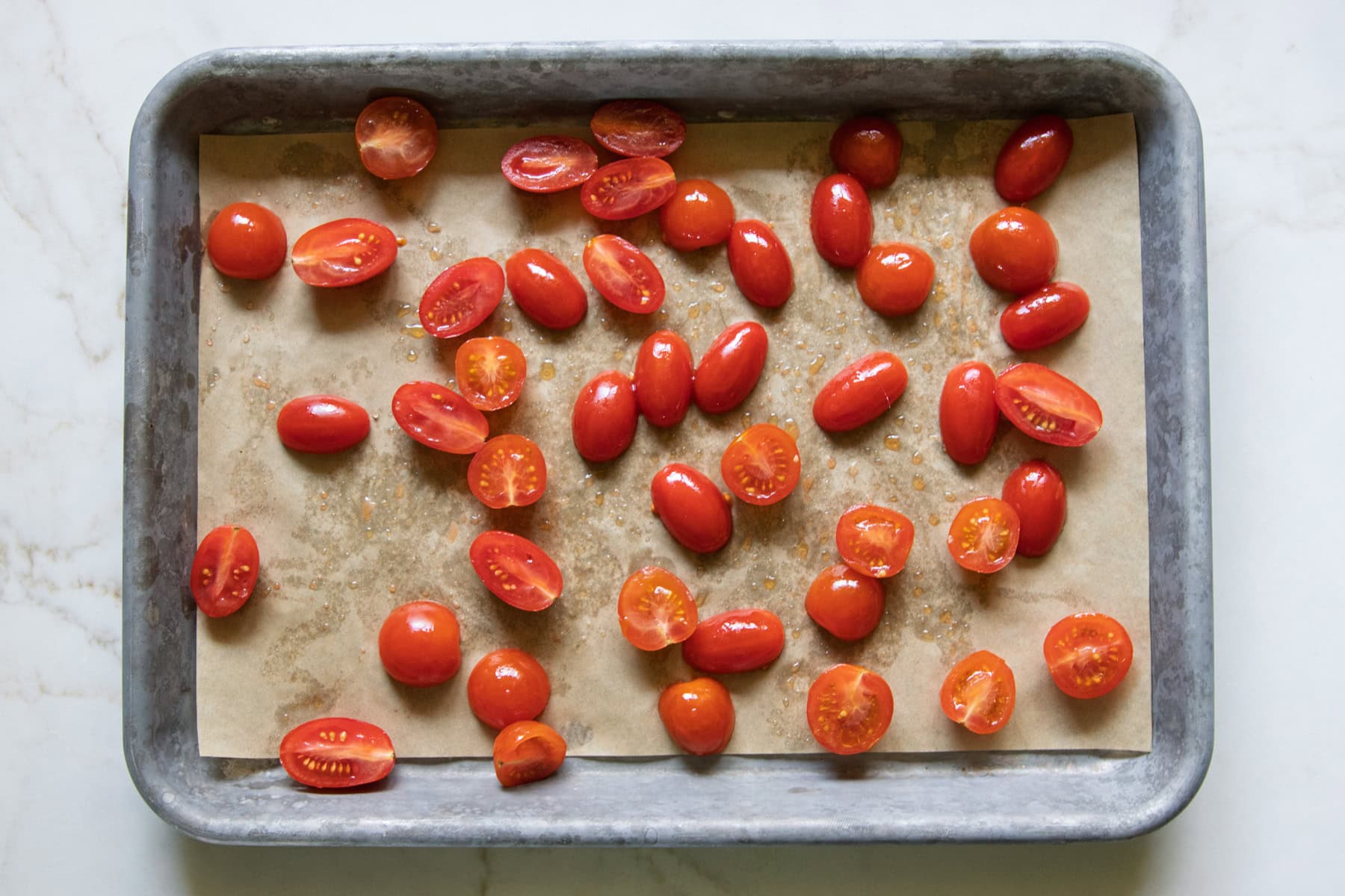 Halved cherry tomatoes tossed in olive oil and spread out on a parchment lined sheet pan ready for roasting.