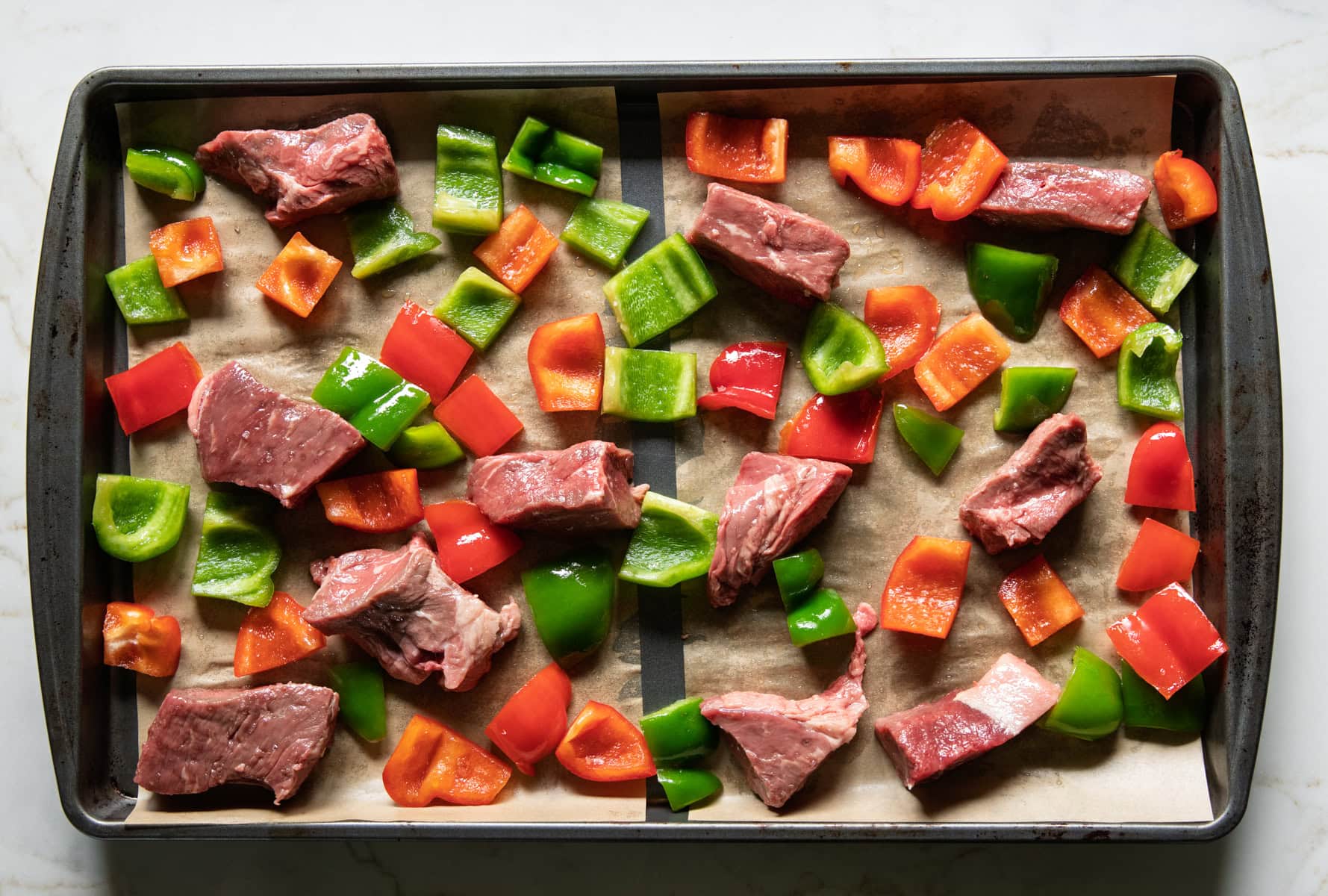 A sheet pan filled with large chunks of top sirloin, red bell pepper, and green bell pepper ready for baking.