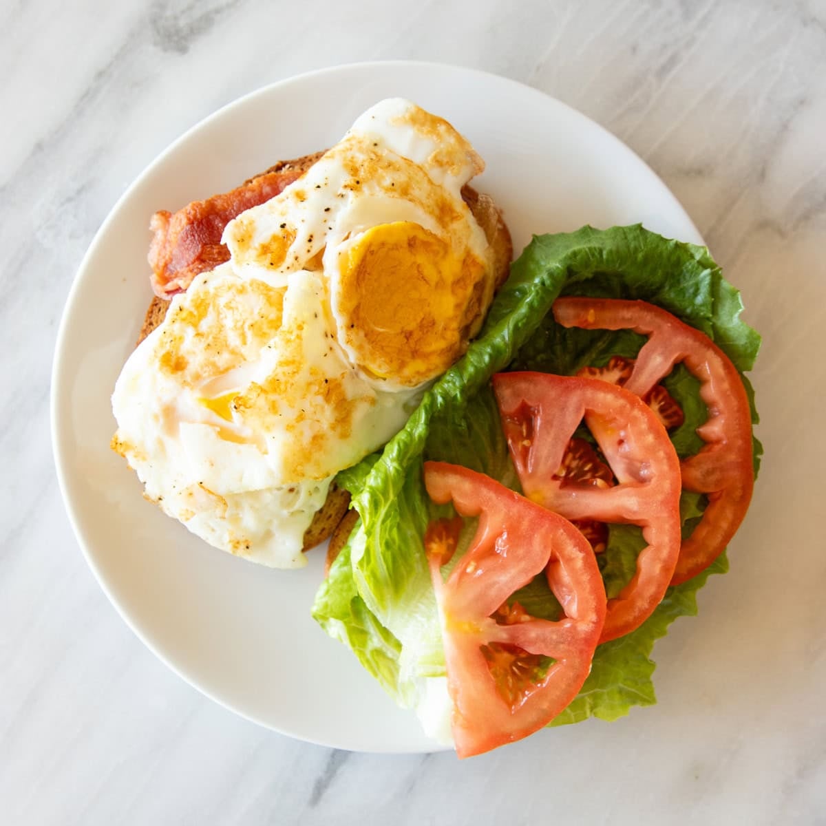 An open-face BLT sandwich. One side has bacon and fried eggs. The other has romaine lettuce and tomato.